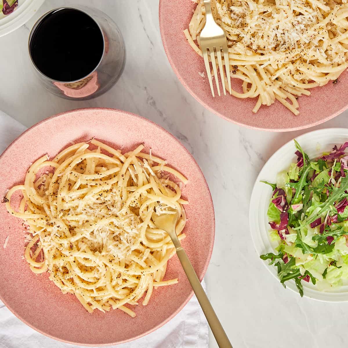 two bowls of bucatini cacio e pepe topped with cracked pepper and grated cheese served with a glass of wine and salad