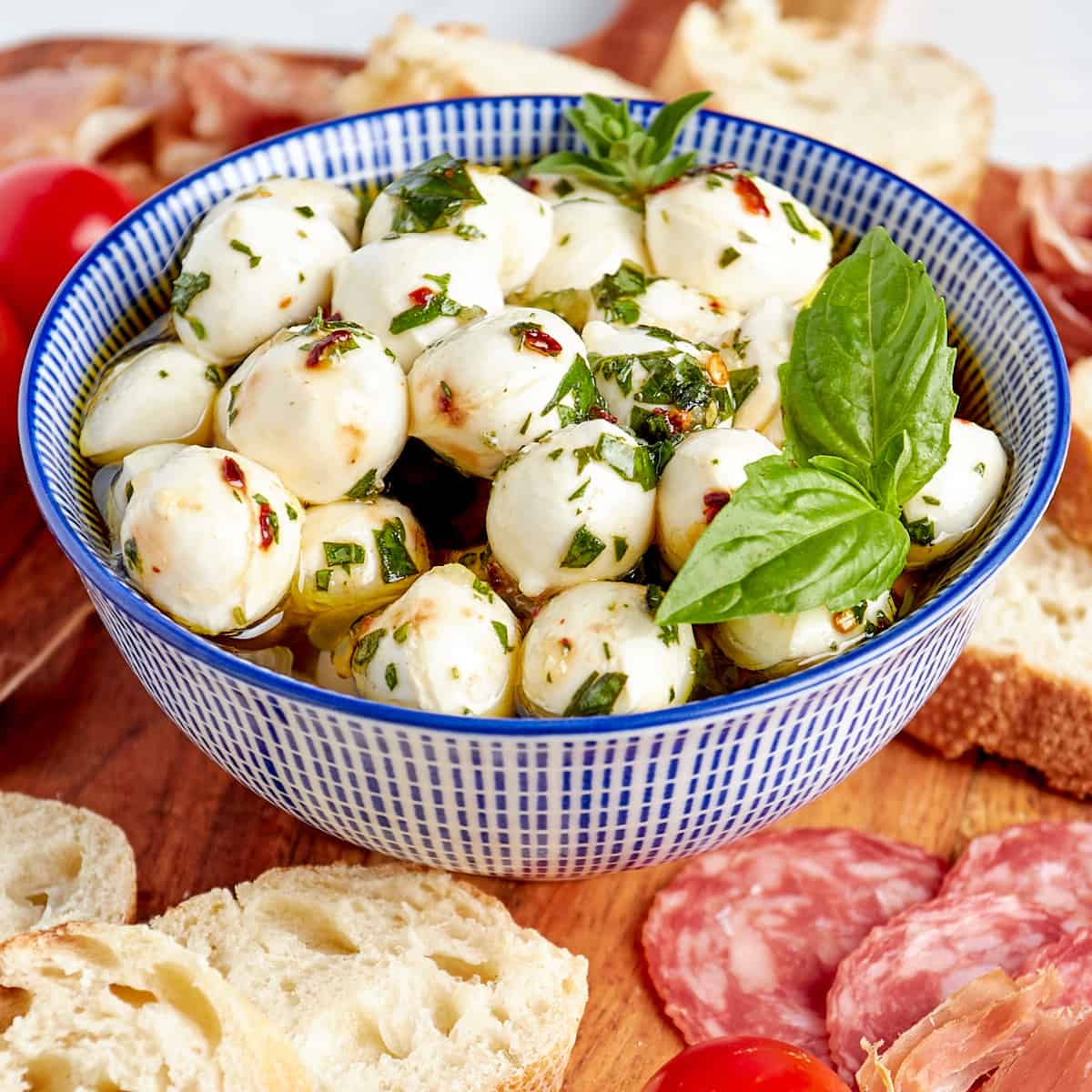 marinated mozzarella balls in a bowl next to bread, cured meat, and tomatoes