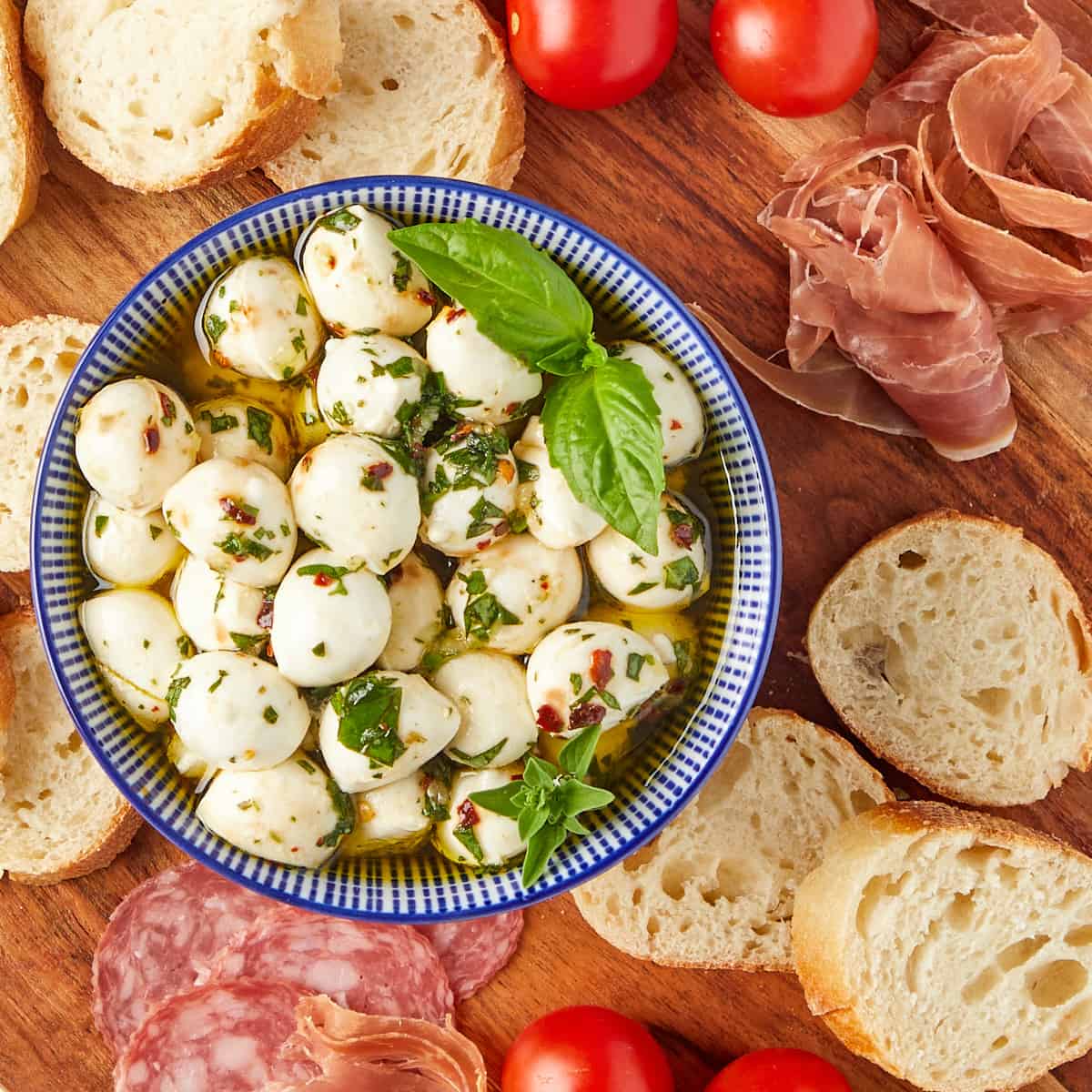 marinated mozzarella balls in a bowl next to bread, cured meat, and tomatoes