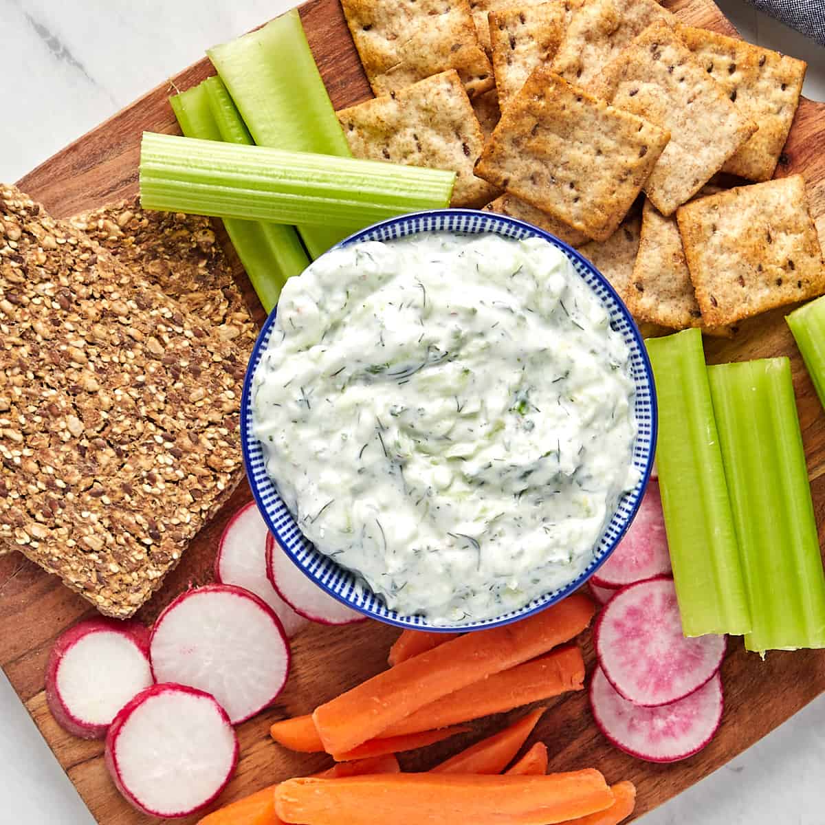 tzatziki sauce in bowl served on a wood board with sliced radishes, carrot sticks, celery sticks, pita crackers, and nut crackers