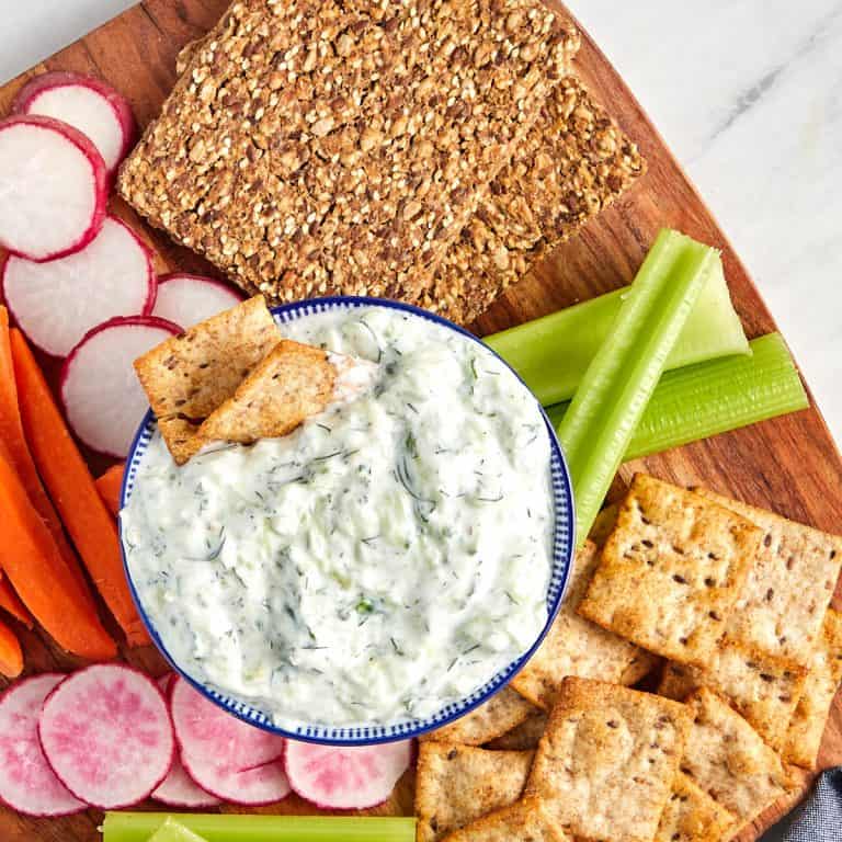 tzatziki sauce in bowl served on a wood board with sliced radishes, carrot sticks, celery sticks, pita crackers, and nut crackers