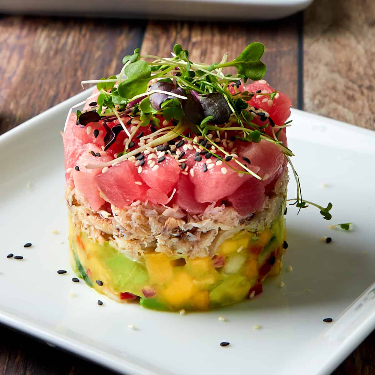 crab and ahi tuna tower on a white plate with a sprinkle of black and white sesame seeds and topped with microgreens