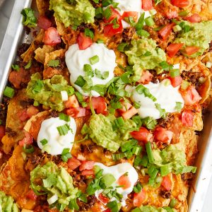 cooked nachos in sheet pan topped with sour cream, tomatoes, guacamole, and green onion