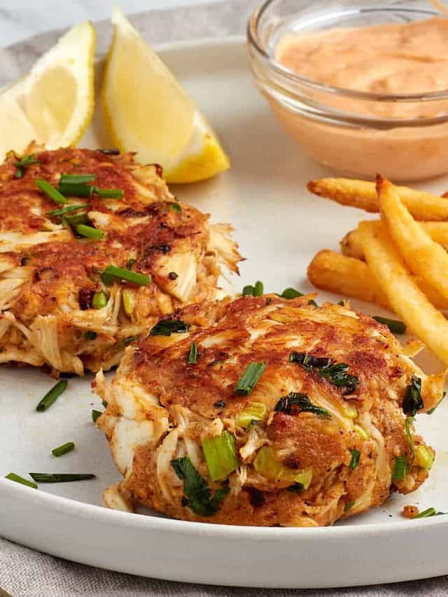 2 crab cakes on a plate with spicy remoulade, fries, and lemon wedges