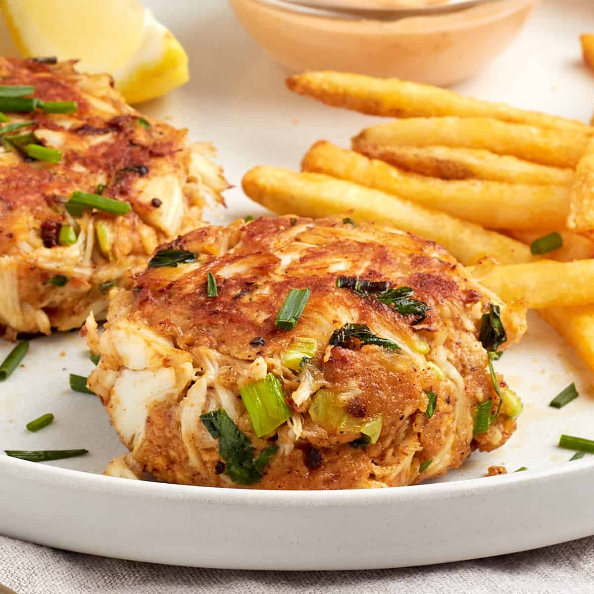 2 crab cakes on a plate with spicy remoulade, fries, and lemon wedges
