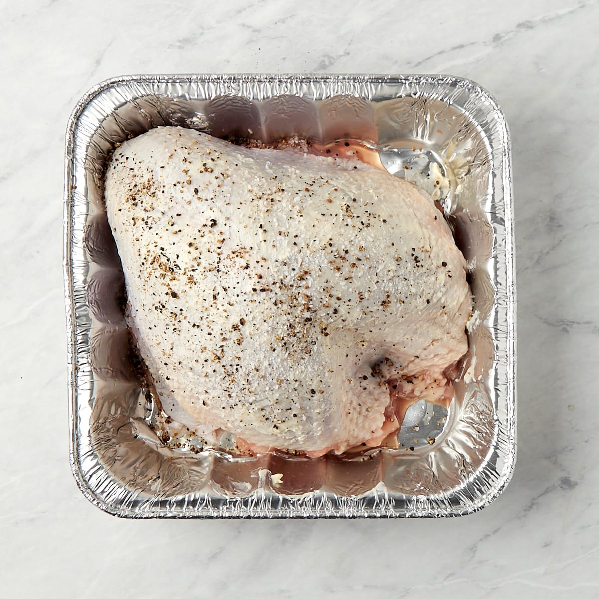 raw turkey breast in an aluminum pan after butter has been rubbed on the skin and seasoned with salt and pepper.