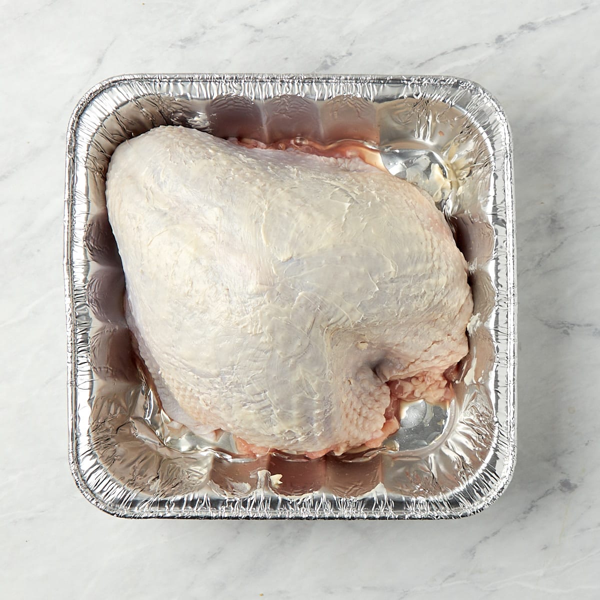 raw turkey breast in an aluminum pan after butter has been rubbed on the skin.