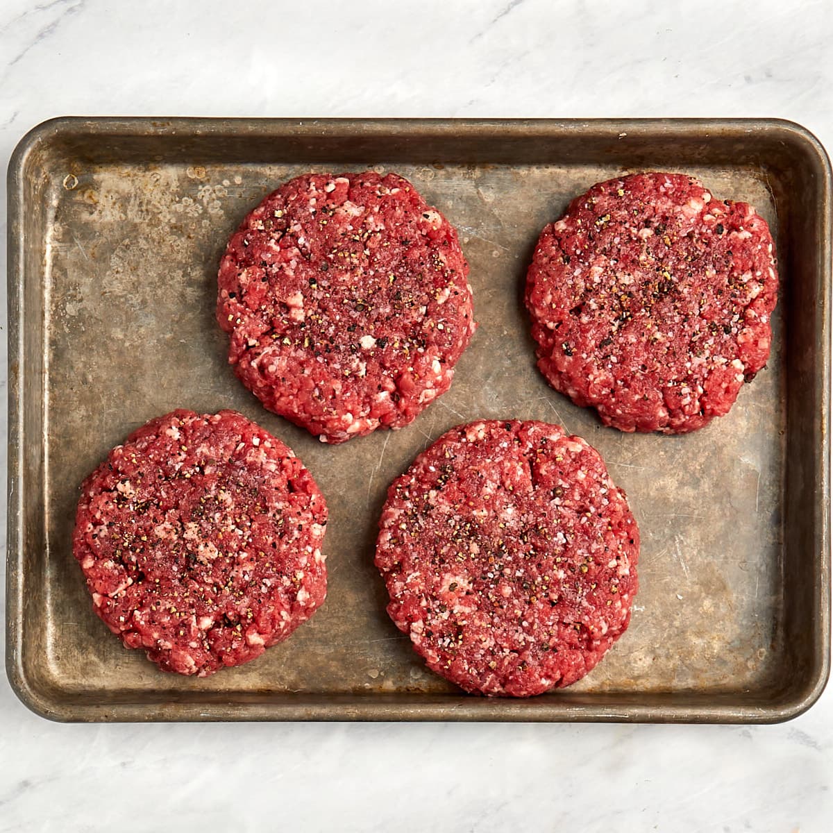 4 burger patties with salt and pepper on a baking sheet.