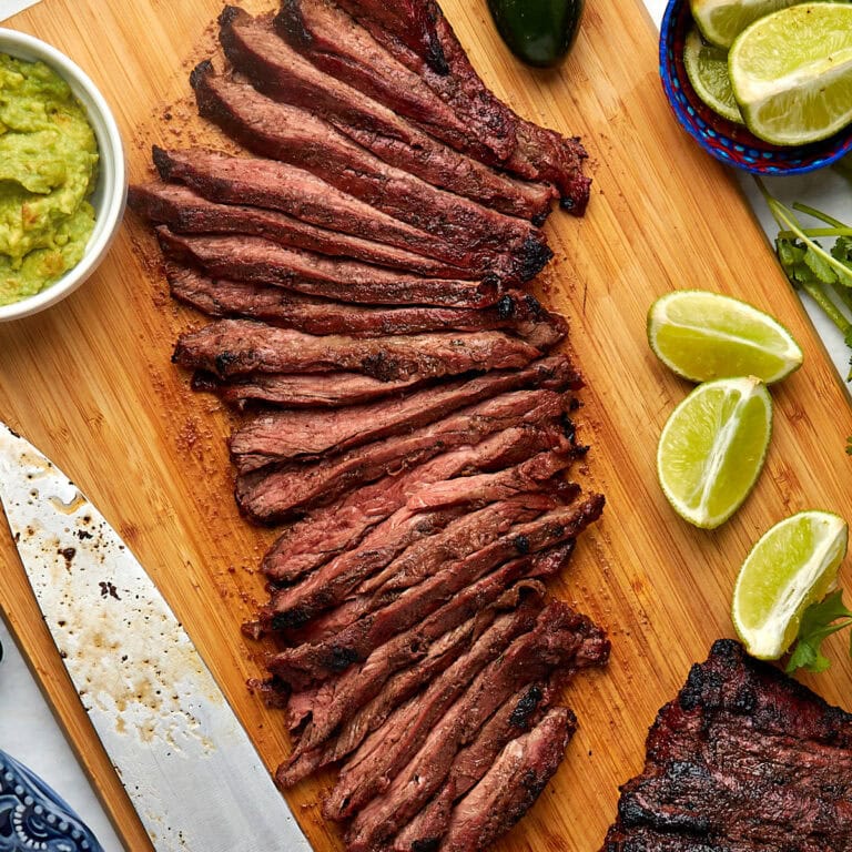 sliced carne asada on a wood cutting board with limes and guacamole on the side.