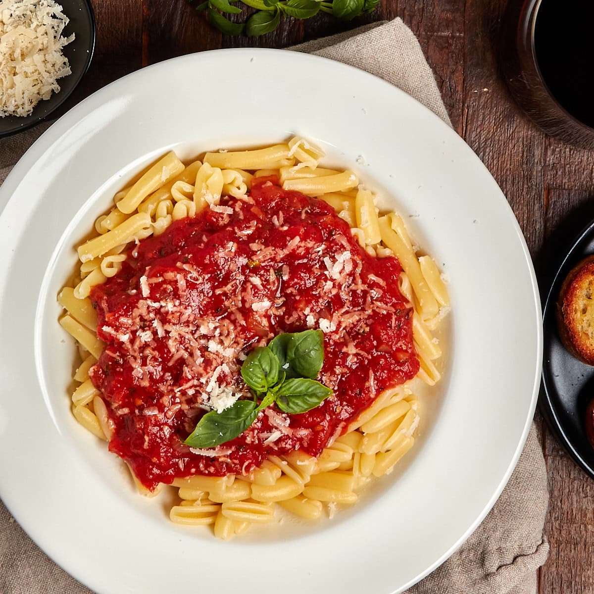a white bowl filled with pasta and marinara sauce topped with basil and grated cheese.