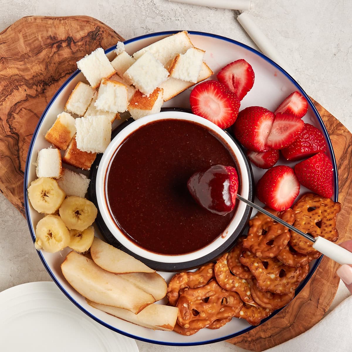 a strawberry being dipped in chocolate sauce in a bowl on a plate with strawberry, banana, pretzels, and angle food cake.