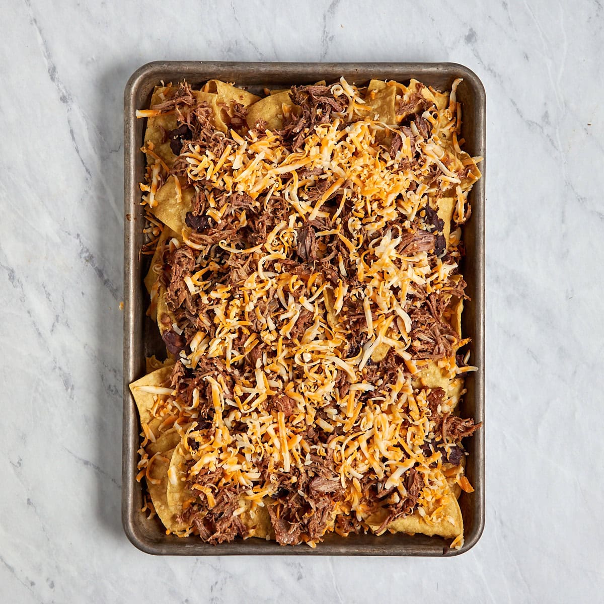 tortilla chips on a sheet pan topped with pulled pork, shredded cheese, and refried beans.