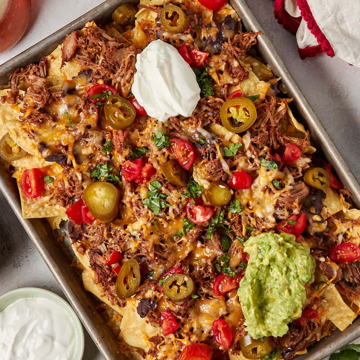 nachos with pulled pork, refried beans, cheese, sour cream, guacamole, tomatoes, and jalapenos on a sheet pan.