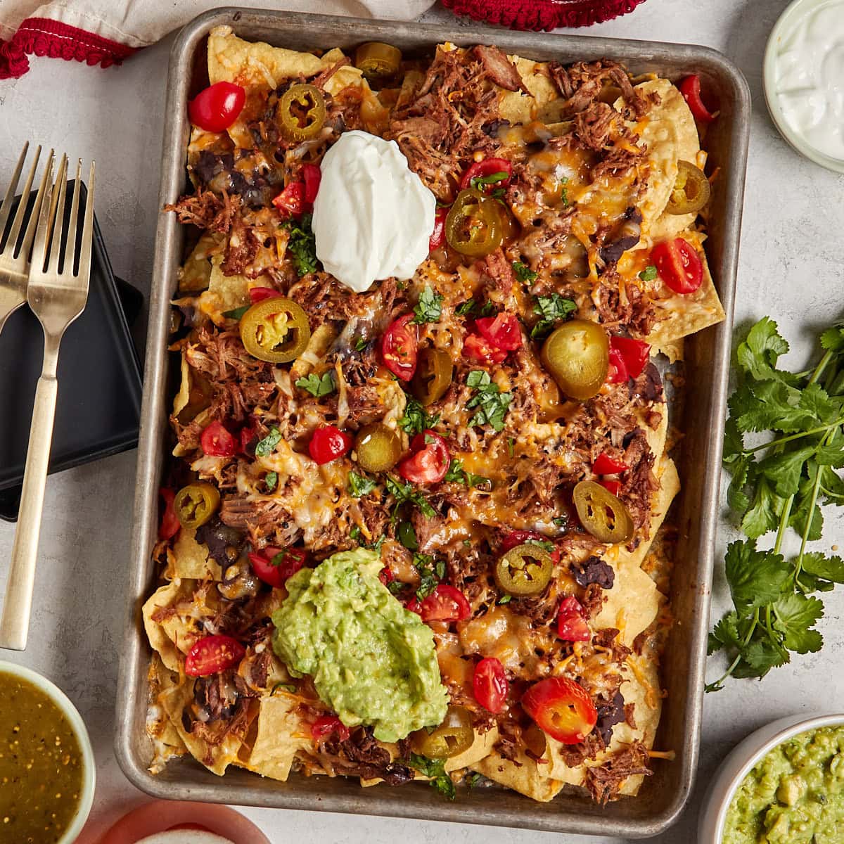 nachos with pulled pork, refried beans, cheese, sour cream, guacamole, tomatoes, and jalapenos on a sheet pan.