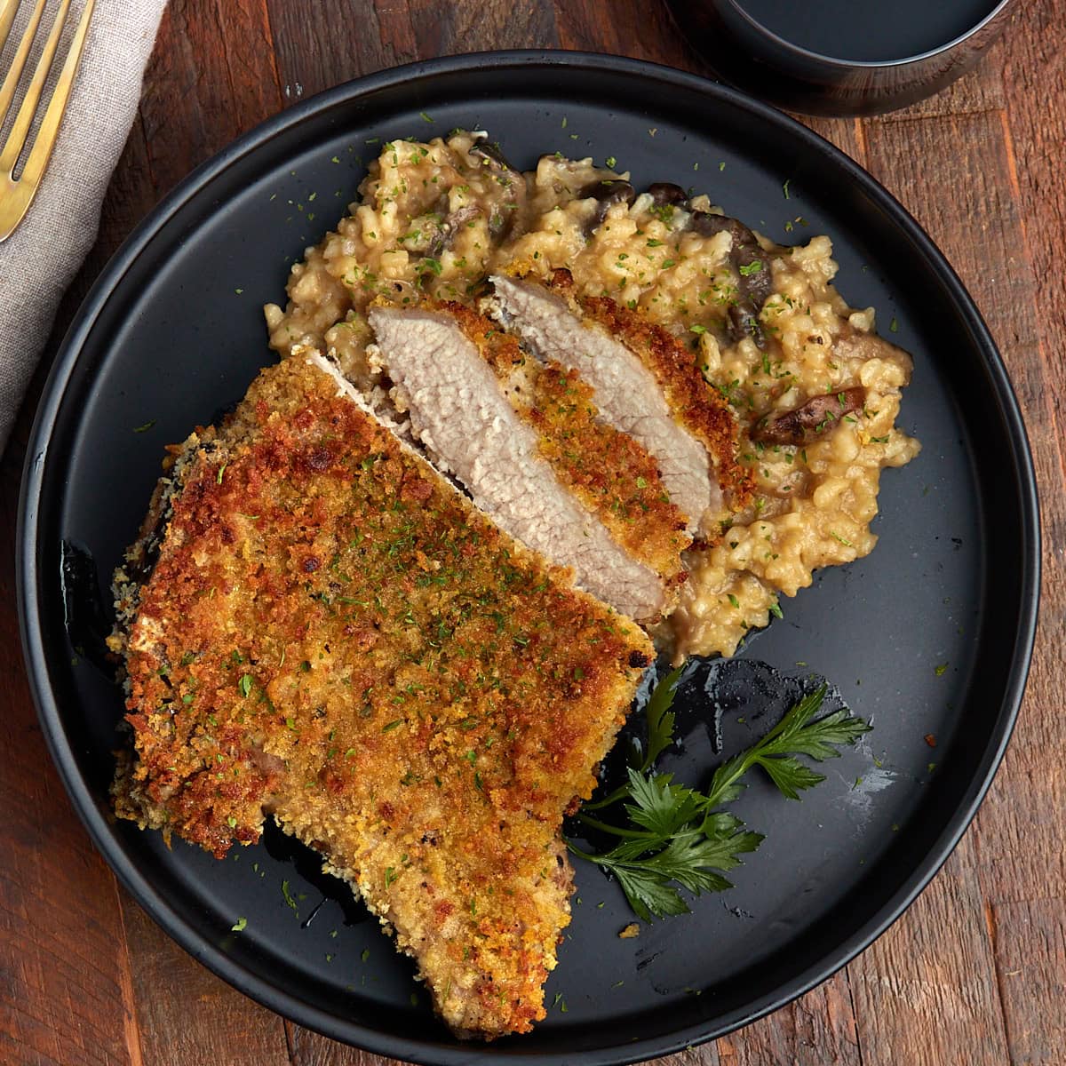 cut breaded bone in pork chops with mushroom risotto on a black plate.