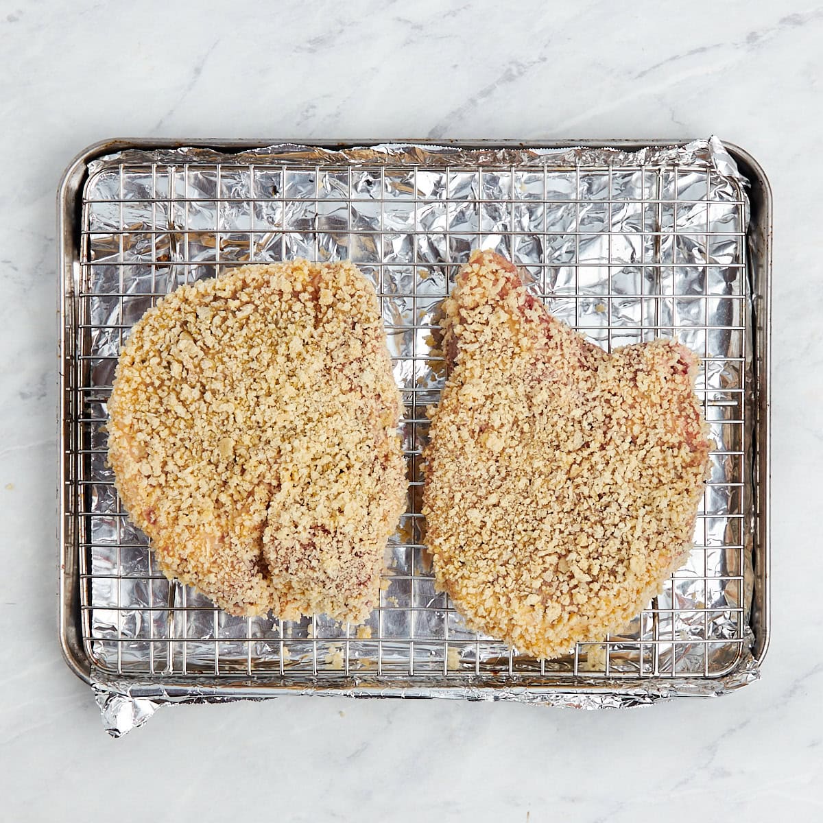 two breaded uncooked bone in pork chops on a wire rack.