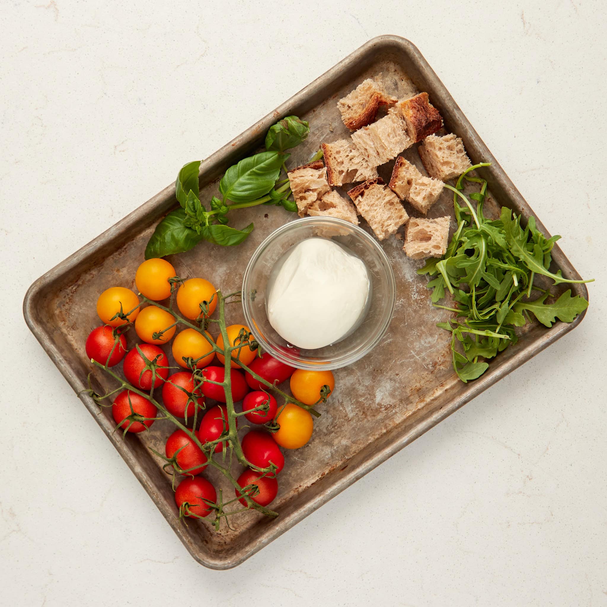 Cherry tomatoes, bread cubes, fresh greens, and yogurt on a rustic tray.