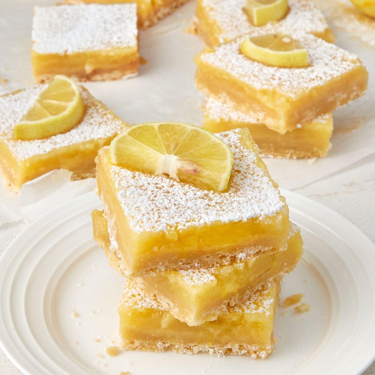 3 lemon bars stacked on top of each other on a white plate with a lemon wedge garnish.