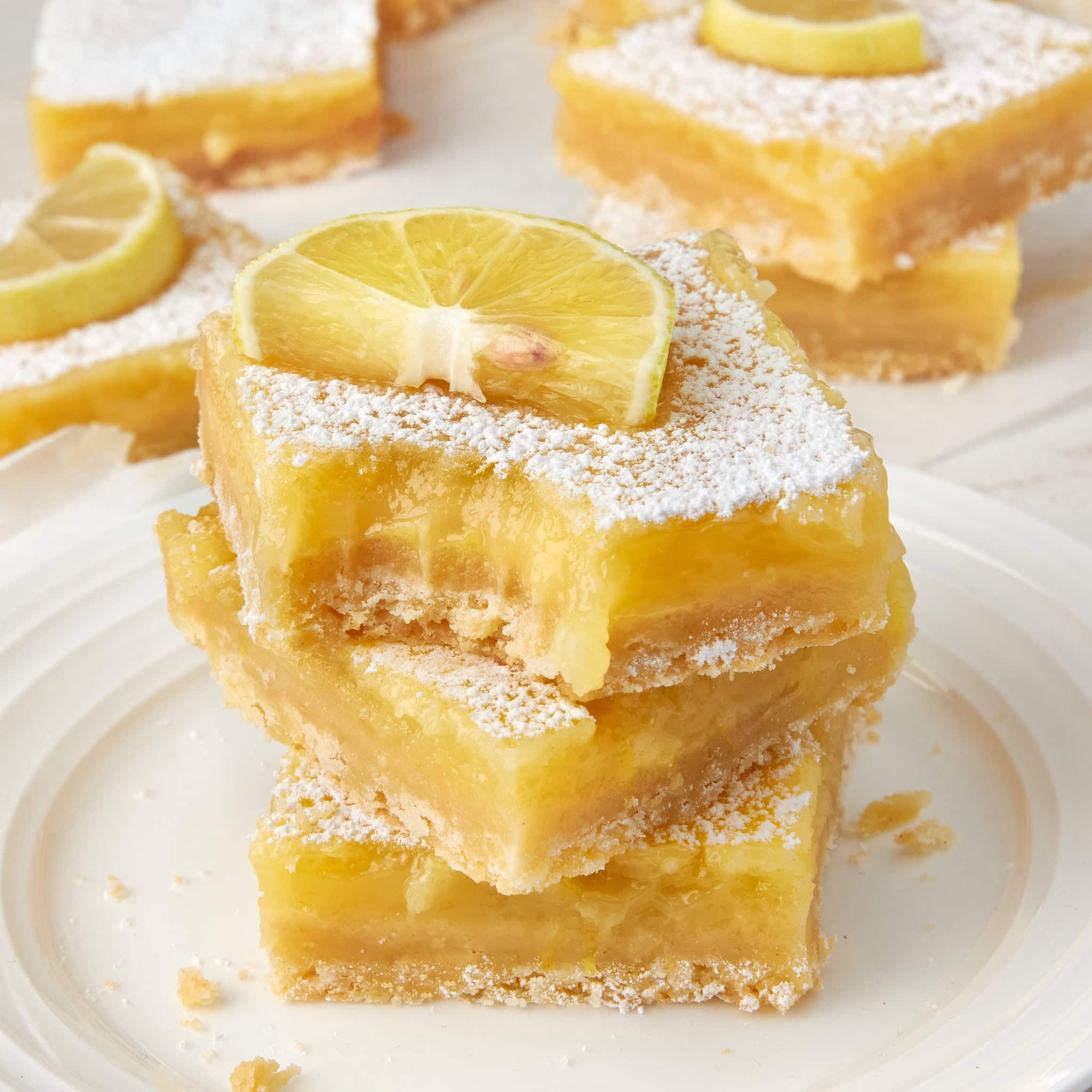 3 lemon bars stacked on top of each other on a white plate with a lemon wedge garnish. the top lemon bar has a bite taken out of it.