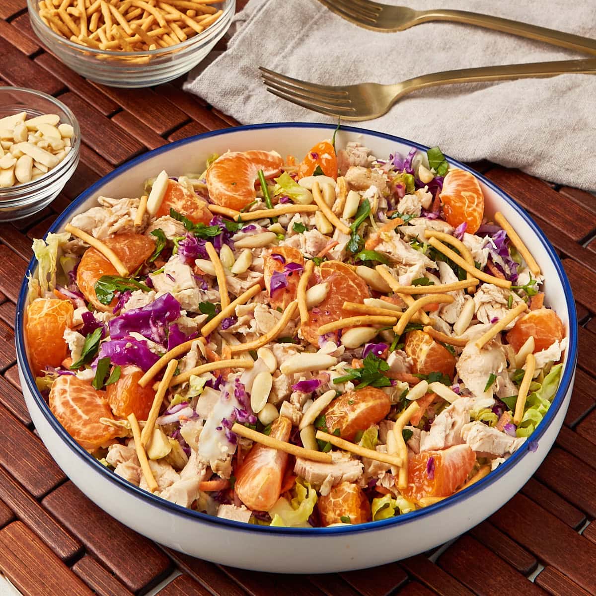 mandarin orange chicken salad served in a white bowl with a side of slivered almonds and chow mein noodles.