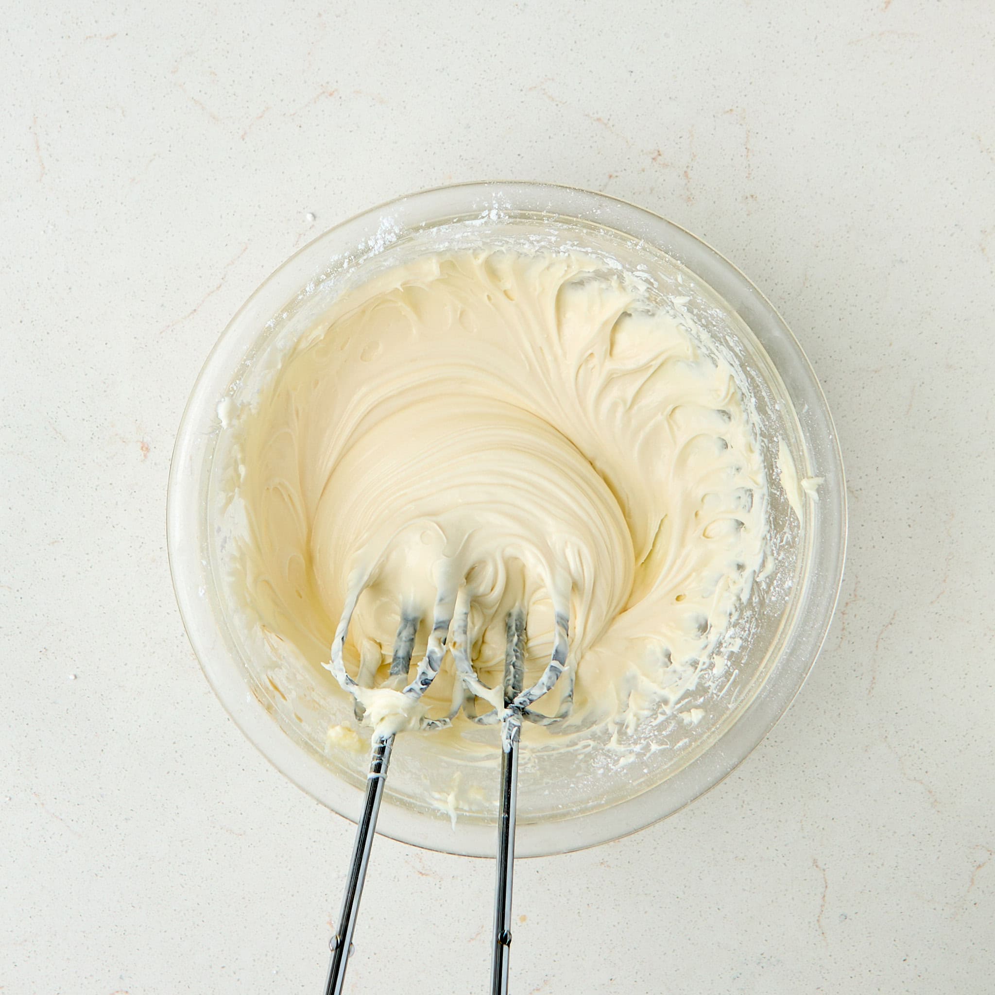 cream cheese frosting in a clear bowl with beaters from a hand mixer.