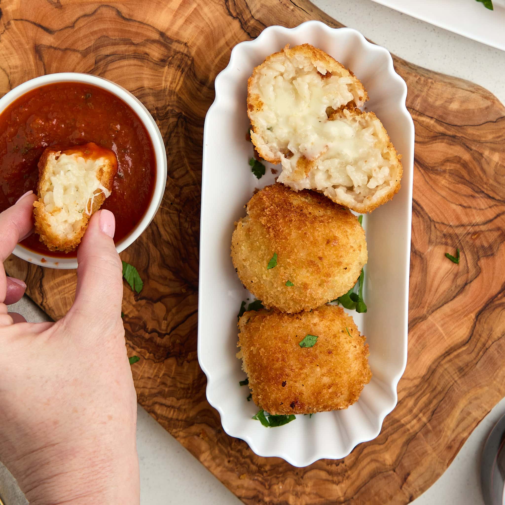 3 arancini in white platter and a piece of one being dipped in marinara sauce.