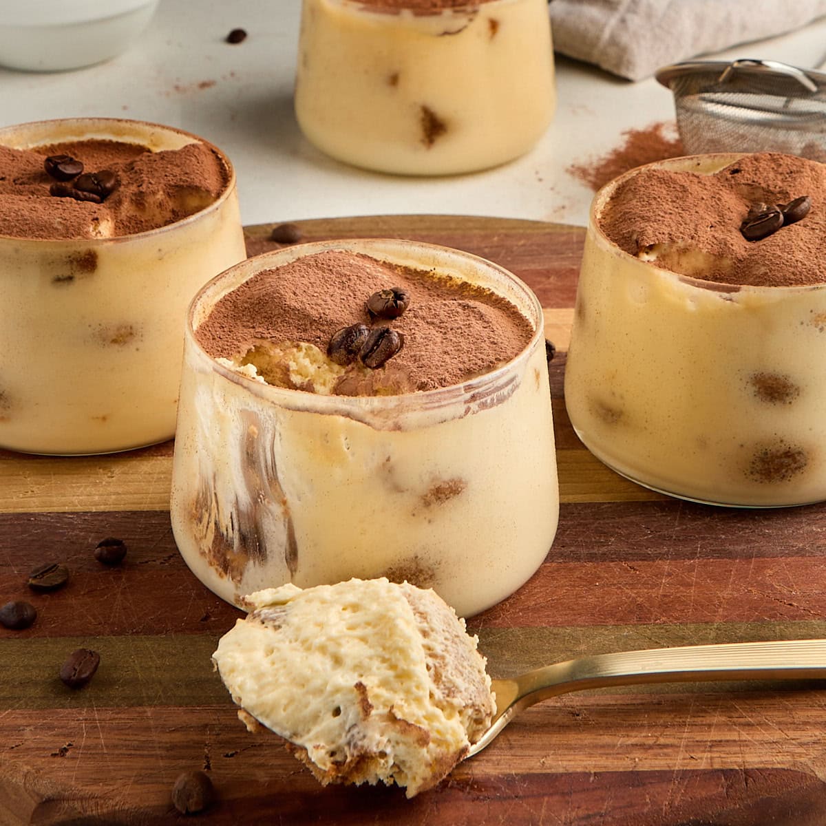 3 tiramisu cups and a spoonful of tiramisu sitting on a wooden board.