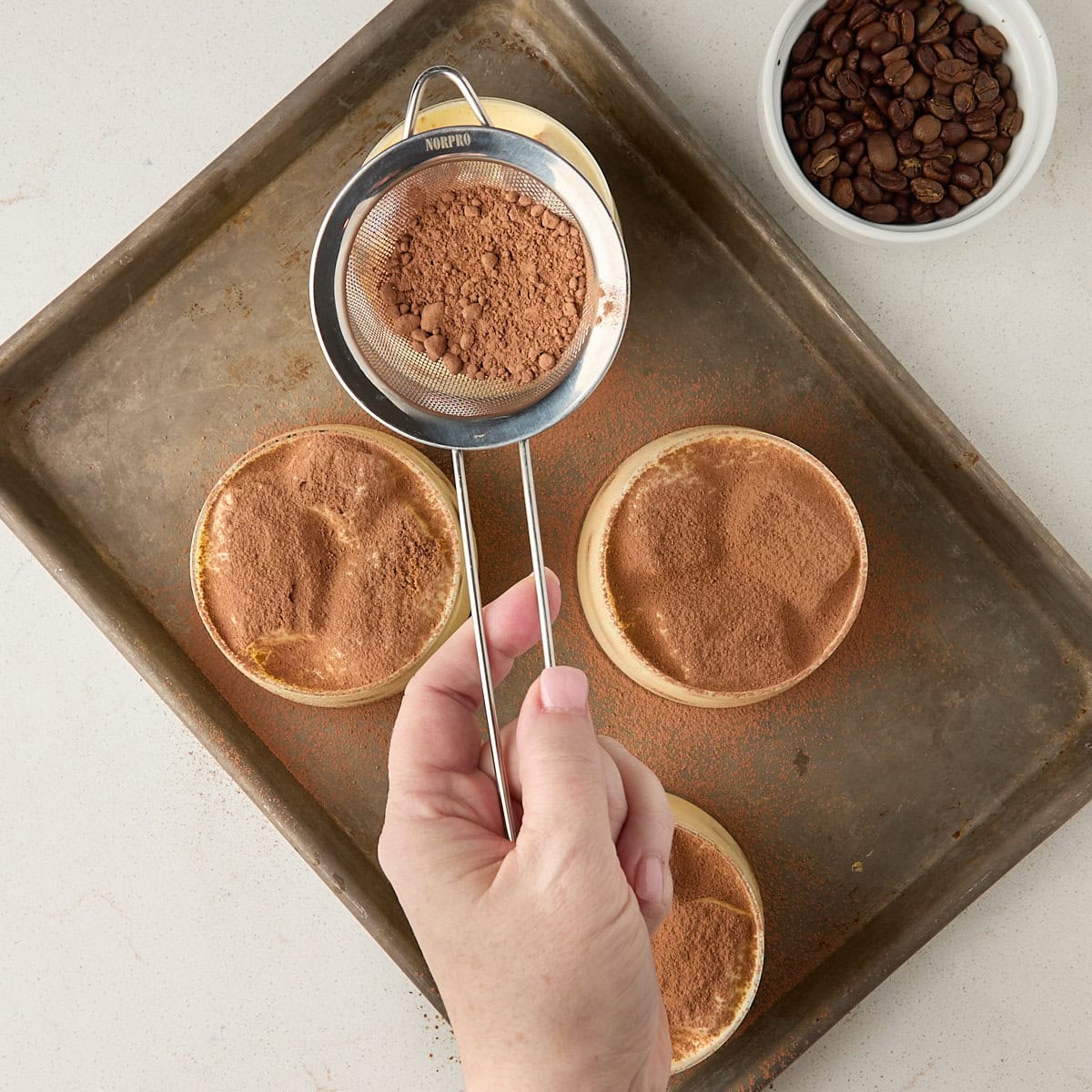 tiramisu cups on a baking sheet being dusted with cocoa powder.